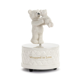 Front view of a white rotating music box. The base has a diamond pattern on the sides. The words "wrapped in love" are printed in gold on the base. There are two teddy bear figures, parent and child, on top. The parent bear is holding the child bear up, so they are face to face