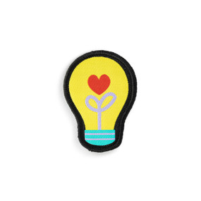 Front view of a light bulb shaped badge in packaging. The badge is a yellow light bulb. The bulb's filaments resemble a flower stem with a red heart as the flower. The badge can be attached to a Just Add Heart Bear.