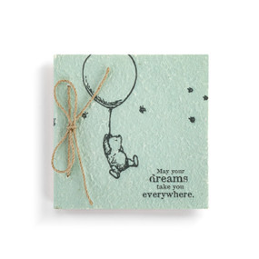 Top down view of the front cover of a green paper mâché journal. The cover has an illustration of classic Winnie-the-Pooh holding on to a balloon. "May your dreams take you everywhere" is printed in the lower right corner. The journal is bound with twine on its left side.