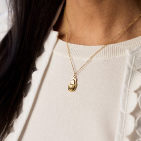 A woman wears a gold necklace with a small charm pendant. The charm is two palm-up hands, one atop the other, with a small heart at the center of the smaller hand's palm.