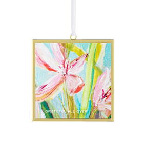 Front view of a glass suncatcher in a gold frame with a white cord threaded through a loop hanger on the top. On the surface of the glass is print of a pink flower and green leaves on a sky blue background. "Grateful all over" is printed on the upper right corner.