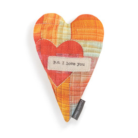 Top down view of an orange and red heart shaped pillow. A large red heart is at the center left, and natural colored patch with the phrase  "p.s. I love you" in typeface is at the center.