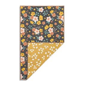 Top down view of the front of a kitchen towel, which has a pattern of pink, orange and yellow flowers on a dark brown background. The bottom right corner is folded up to show the back of the towel, which has a print of white flowers on a mustard yellow background.