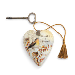 Top down view of a beige and gold art heart. The front of the heart has a design of  brown and yellow songbird sitting in fall foliage on a white and beige background. The phrase "Thankful and Blessed" is printed in script on the heart's upper left side. A gold cord with a tassel at the end is attached to the top and displayed to the heart's left. The cord is threaded through a bronze key that can be attached at the back as a stand.