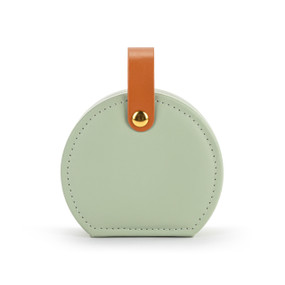 Front facing view of a circular sage green traveling jewelry case. The brown strap is snapped at the top, holding the case closed.