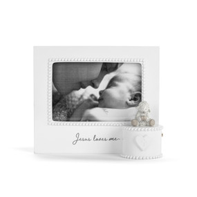 Front view of a white musical frame. The frame's opening has a white bead-like border. The music box mechanism is attached to the lower left corner of the front of the frame in a white circular box, and has a figure of a lamb on top and a heart with a cross at the center in front. The phrase "Jesus Loves Me" is printed in black script under the frame's opening.
