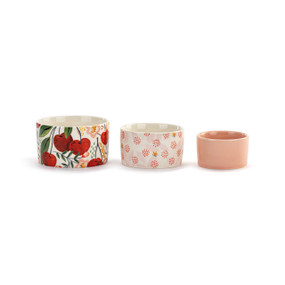 Front view of three nesting prep bowls. The bowls are set in a line, with the largest on the left and the smallest on the right. The largest bowl has a pattern of cherries on stems with leaves. The middle sized bowl has a pattern of small red dots clustered together to look like cherries. The smallest bowl is pink.