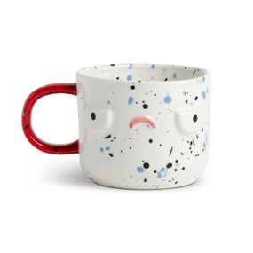 Front facing view of a white character mug that has green and blue speckles. The mug's red handle is to the left. The mug is named Nico Morning Grump and has a grumpy face on his front.