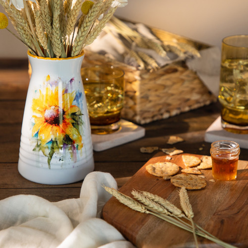 A white ceramic vase with a watercolor image of a yellow sunflower on it sitting on a wood table surrounded by snacks and drinks.