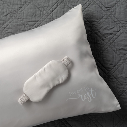 light gray satin eye mask with wide elastic band on top of light gray satin pillow, both with the phrase 'invest in rest' in the bottom right corner