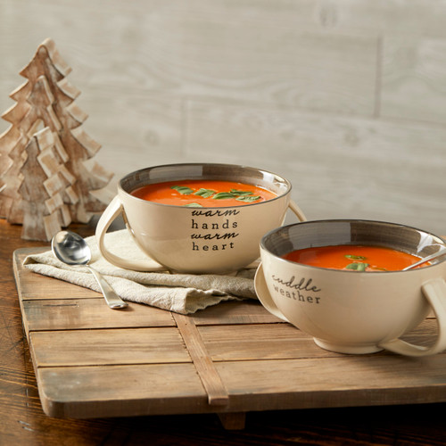 two cream colored ceramic bowls reading Warm Hands Warm Heart and Cuddle Weather filled with tomato soup set on a table