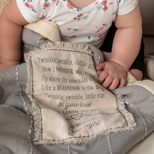 A close up image of a baby girl, sitting on an ivory couch, snuggled in a gray and ivory blanket with a large patch that reads 'Twinkle Twinkle Little Star'.