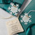 A set of two snowflake shaped ornaments that say "Always Sisters" with a cream drawstring bag with a message about one to keep and one to share.