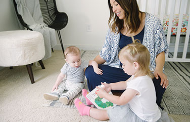 A mom sitting on the ground with two blonde babies, a chair, and a crib