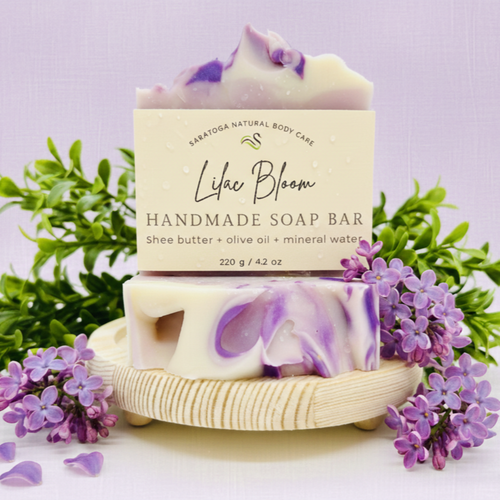 Handmade lilac soap bar with purple and cream swirl, wrapped in Saratoga Natural Body Care label, styled with fresh lilac flowers on a wooden soap dish