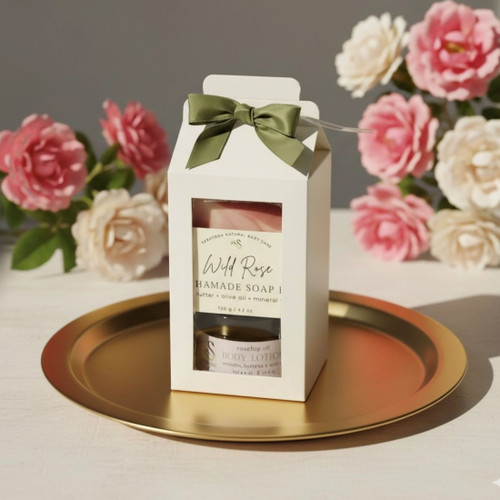 Single Gift Set on a Gold Tray. A single white gift box is centered on a shiny gold round tray. The box, tied with a green ribbon, has a clear window revealing the "Wild Rose" soap and lotion. The background is a cluster of blurred pink and white roses, highlighting the product's floral scent. Single Gift Set on a Gold Tray. A single white gift box is centered on a shiny gold round tray. The box, tied with a green ribbon, has a clear window revealing the "Wild Rose" soap and lotion. The background is a cluster of blurred pink and white roses, highlighting the product's floral scent.