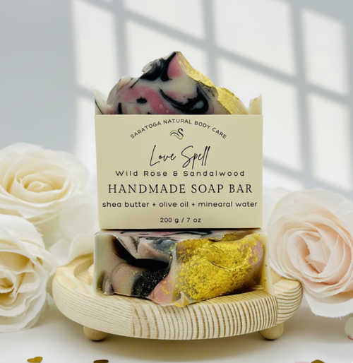 Handmade Love Spell soap bar with wild rose and sandalwood scent, pink and black swirl design, displayed on a wooden soap dish with white roses.