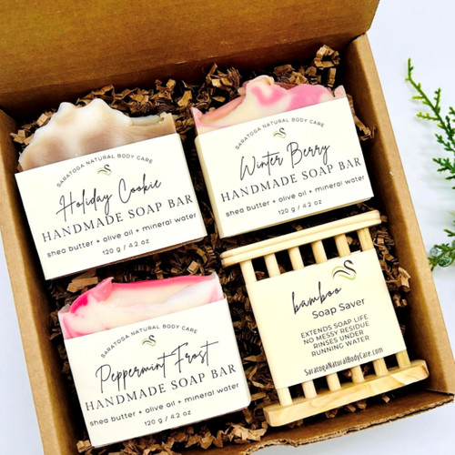 Holiday gift box featuring three handmade soap bars — Peppermint Frost, Winter Berry, and Spiced Cookie — plus a bamboo soap saver. Vegan, botanical, and beautifully packaged holiday skincare gift set.