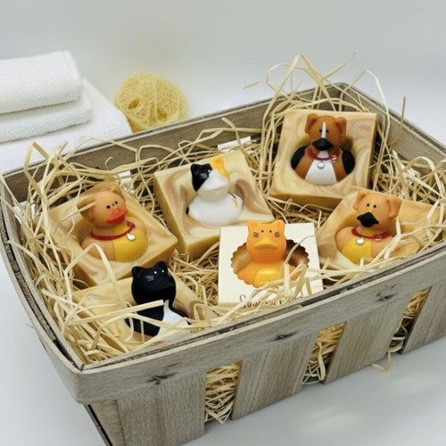 A top-down view of a light-colored wooden basket filled with five bars of Soapy Pals soap and straw-like packing material. The soaps feature various pet-themed rubber duckies, including a black-and-white cat, a calico cat, two brown dogs, and an orange cat in its paper packaging. In the background, there are folded white towels and a loofah. A top-down view of a light-colored wooden basket filled with five bars of Soapy Pals soap and straw-like packing material. The soaps feature various pet-themed rubber duckies, including a black-and-white cat, a calico cat, two brown dogs, and an orange cat in its paper packaging. In the background, there are folded white towels and a loofah.