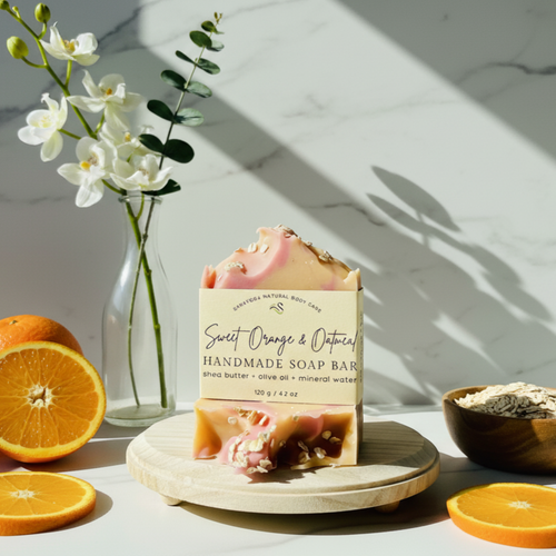 Handmade Sweet Orange and Oatmeal soap bar with shea butter, olive oil, and mineral water, displayed on a wooden stand beside a glass vase with white flowers in natural sunlight. Handmade Sweet Orange and Oatmeal soap bar with shea butter, olive oil, and mineral water, displayed on a wooden stand beside a glass vase with white flowers in natural sunlight.
