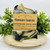 Mahogany Charcoal handmade soap bar by Saratoga Natural Body Care, featuring black and cream swirls, displayed on a wooden stand with greenery accents.