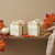 A pair of handcrafted soap bars, "Crisp Apple Sage" and "Pumpkin Sandalwood", are presented on a wooden table with fall-themed decorations. The "Crisp Apple Sage" bar is a marbled mix of green, pink, and white, while the "Pumpkin Sandalwood" is a solid cream color. A small bundle of cinnamon sticks and scattered maple leaves sit in front of the soaps. A pair of handcrafted soap bars, "Crisp Apple Sage" and "Pumpkin Sandalwood", are presented on a wooden table with fall-themed decorations. The "Crisp Apple Sage" bar is a marbled mix of green, pink, and white, while the "Pumpkin Sandalwood" is a solid cream color. A small bundle of cinnamon sticks and scattered maple leaves sit in front of the soaps.