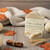 "Pumpkin Sandalwood" Soap Bar. A solid, creamy beige-colored handcrafted soap bar, also stacked and placed on a wooden surface with a soft, knit blanket behind it. The label reads "Saratoga Natural Body Care," "Pumpkin Sandalwood Handmade Soap Bar," and specifies the same ingredients and weight. The surrounding decorations are similar, featuring scattered autumn leaves, cinnamon sticks tied with twine, and white pumpkins. "Pumpkin Sandalwood" Soap Bar. A solid, creamy beige-colored handcrafted soap bar, also stacked and placed on a wooden surface with a soft, knit blanket behind it. The label reads "Saratoga Natural Body Care," "Pumpkin Sandalwood Handmade Soap Bar," and specifies the same ingredients and weight. The surrounding decorations are similar, featuring scattered autumn leaves, cinnamon sticks tied with twine, and white pumpkins.