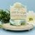 "White Tea & Ginger" Soap Bar. A handmade bar of "White Tea & Ginger" soap from Saratoga Natural Body Care is displayed on a small, round wooden pedestal. The soap features a soft swirl of pale blue and creamy white. An identical, unwrapped bar lies flat underneath. The product is surrounded by sprigs of green foliage and a large white flower. The background is a solid light blue wall. "White Tea & Ginger" Soap Bar. A handmade bar of "White Tea & Ginger" soap from Saratoga Natural Body Care is displayed on a small, round wooden pedestal. The soap features a soft swirl of pale blue and creamy white. An identical, unwrapped bar lies flat underneath. The product is surrounded by sprigs of green foliage and a large white flower. The background is a solid light blue wall.