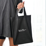 A black tote bag with two handles, featuring a logo on one side, held by a person.