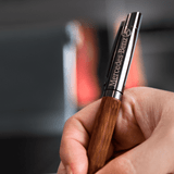 A wooden and metal ballpoint pen with a branding logo, being held in a hand.