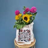 A tote bag in cream fabric displays a floral design and holds an assortment of vibrant flowers, including sunflowers and dahlias.