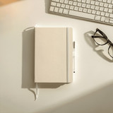 A large hardbound journal in cream with a pen, resting on a desk beside a keyboard and glasses.