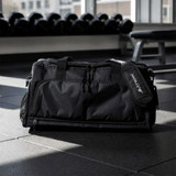 A black duffle bag with multiple zippered compartments and a logo, resting on a gym floor.