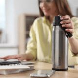 A drink bottle in stainless steel with a black cap, featuring a logo, placed on a desk next to a notebook and phone.