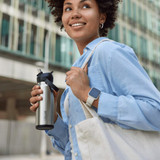 A drink bottle in stainless steel, held by a person wearing a blue shirt and carrying a tote bag. The bottle features a flip straw and has a logo.