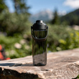 A drink bottle in a sleek, transparent design with a black lid and handle, set against a natural outdoor backdrop.