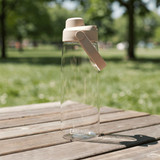 A drink bottle with a clear body and a beige lid, featuring a handle, set on a wooden table in a grassy area.
