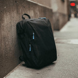 A black travel backpack with blue zipper pulls leaning against a concrete wall. It features multiple compartments.
