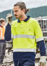 A man wears a bright yellow and navy long sleeve polo with reflective tape, holding a walkie-talkie. It has a logo.