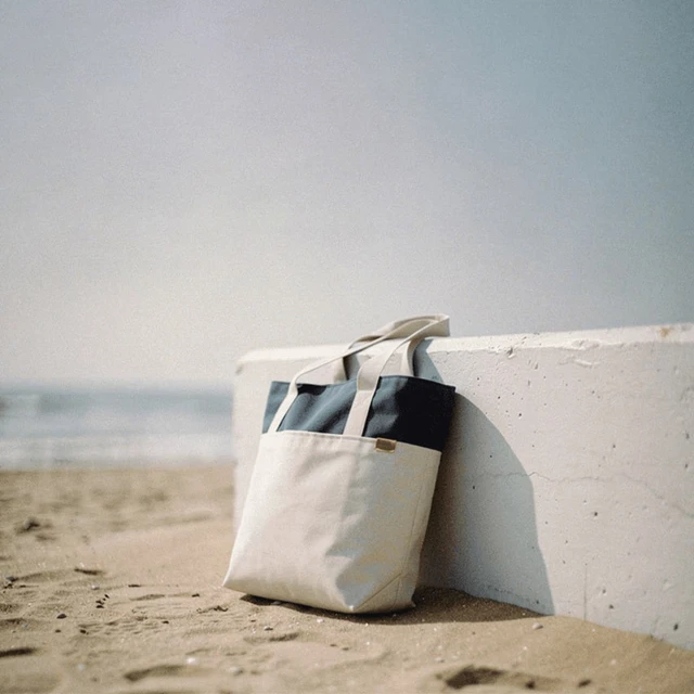 A beach tote bag in cream and navy rests against a white wall on sandy shore, with the ocean in the background.