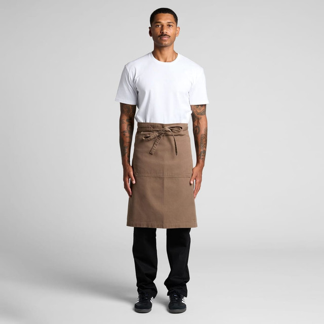 A man wears a brown canvas half apron over a white t-shirt and black pants, with a tied waist and a logo.