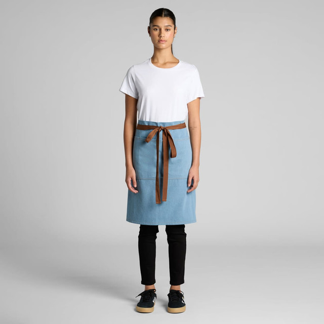 A denim half apron in light blue with a brown tie, worn over a white t-shirt and black pants. Features a logo.