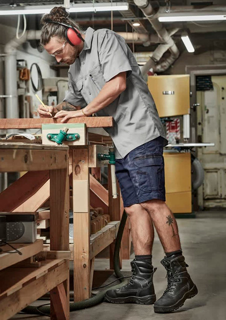 A man wearing grey work shirt and navy shorts stands at a workbench, using a pencil to mark wood. He has ear protection on.