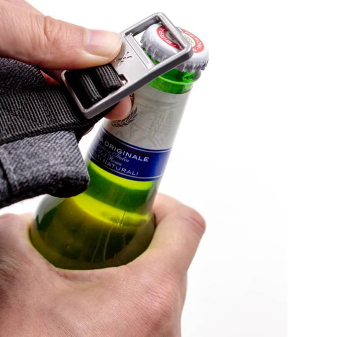 A hand holding a bottle with a grey backpack strap, using a bottle opener attached to the strap.
