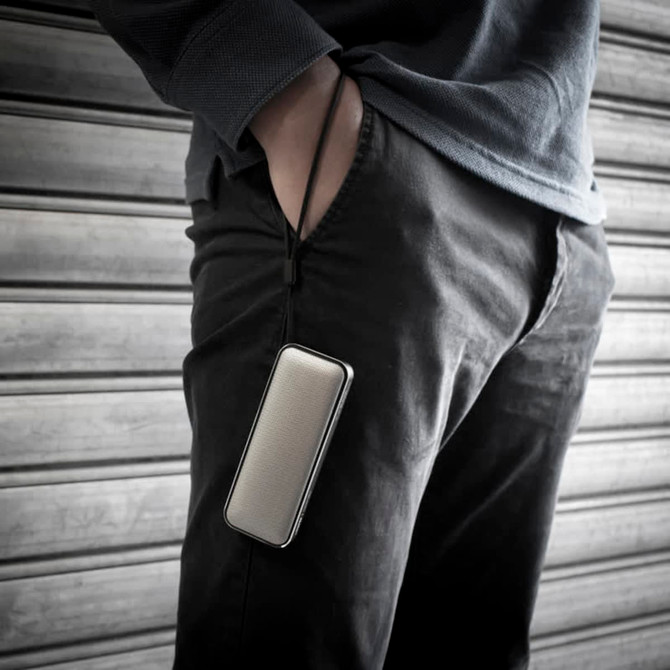 A compact silver Bluetooth speaker is hanging from a person's pocket, featuring a sleek design and a lanyard attachment.