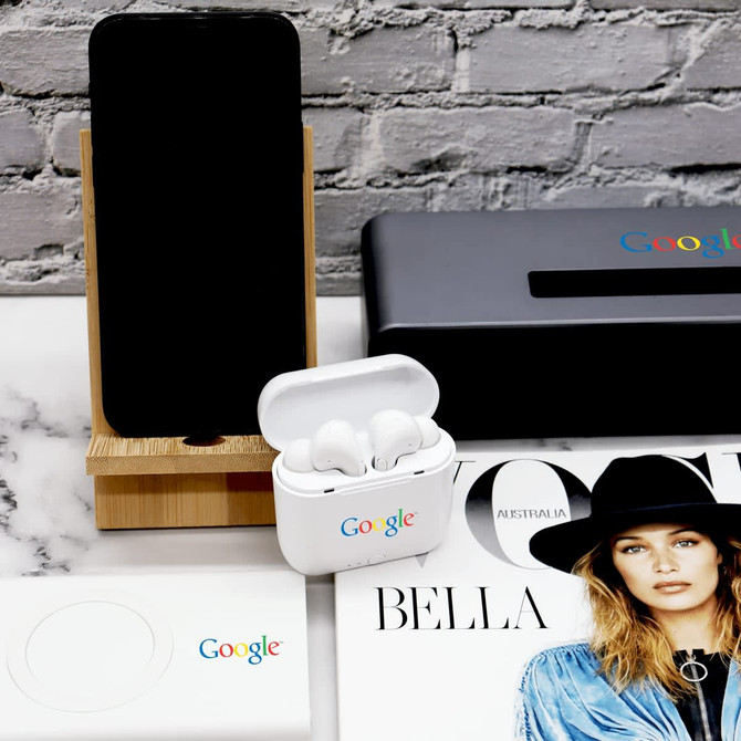 A bamboo phone stand displays a phone, alongside white earbuds in a case and a magazine on a marble surface.