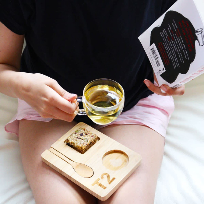 A glass mug with tea, a wooden tray with a spoon, and a treat, held by a person reading a book.