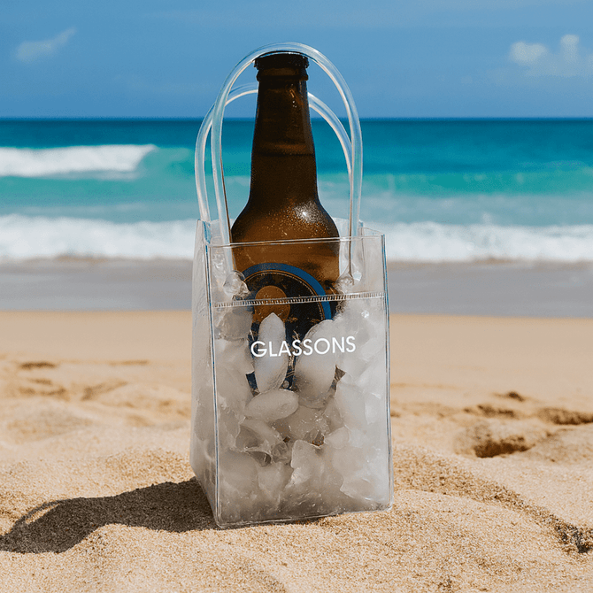 A clear cooler bag filled with ice, holding a brown drink bottle, placed on sandy beach by the ocean. It has a logo.