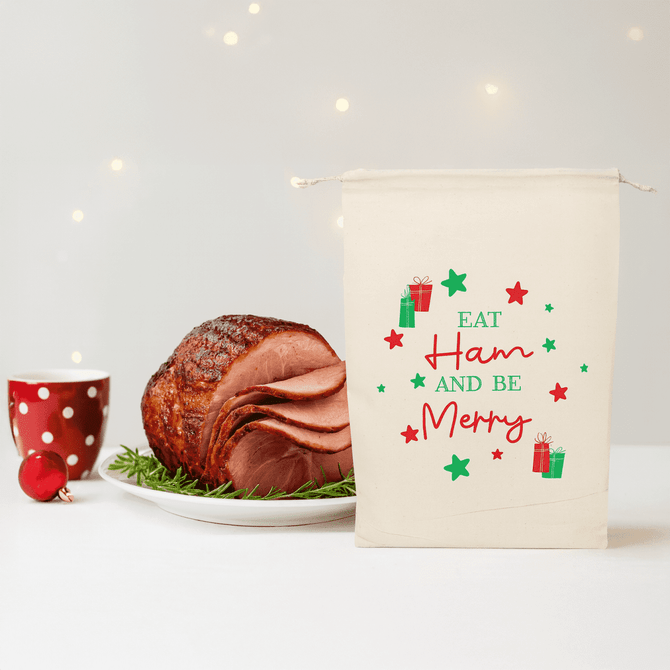A cream-coloured bag with festive designs next to a glazed ham on a platter and a red polka dot cup.