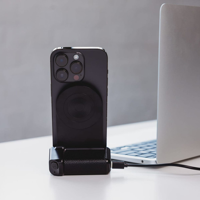 A black wireless charger with a phone attached, placed next to a laptop on a white surface.